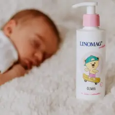 Śpiące dziecko obok butelki oliwki dla dzieci Linomag. Uroczy miś na deskorolce zdobi etykietę.