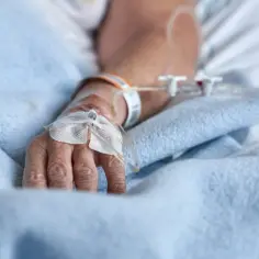 Ręka pacjenta z wenflonem, przez który podawana jest wankomycyna. Dłoń spoczywa na niebieskim kocu.